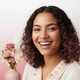 Close-up portrait of a young woman with radiant, clear skin, smiling gently against a soft, light rose and white background.